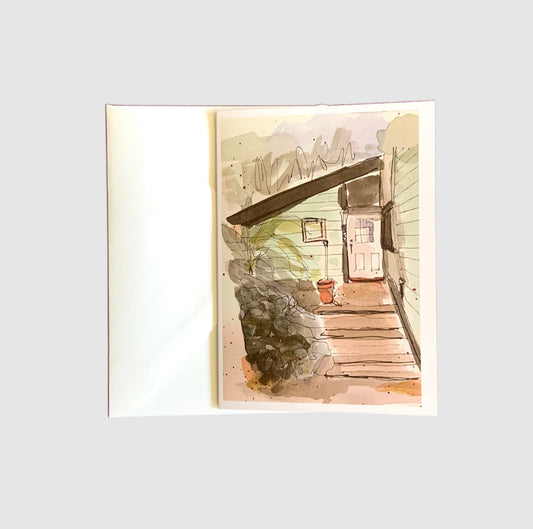 A sketch of a front door surrounded by rocks and trees painted with watercolors on the front of a greeting card.