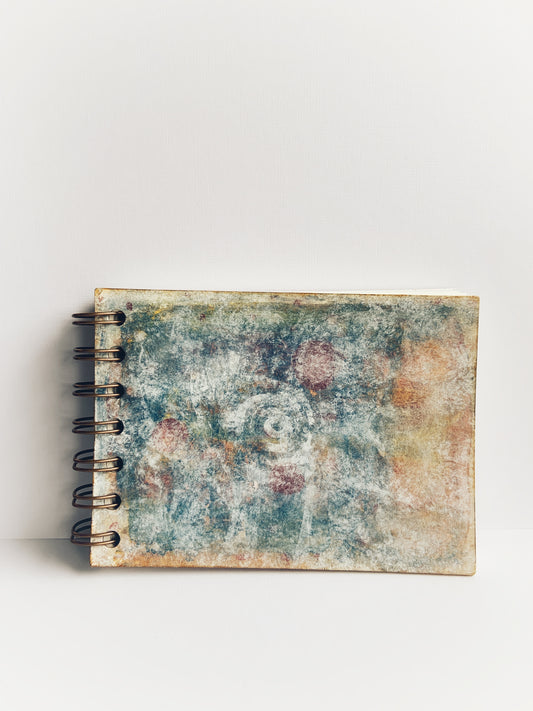 Small coil-bound handmade journal with a faded-white spiral in the centre of a muted blue and orange painted cover