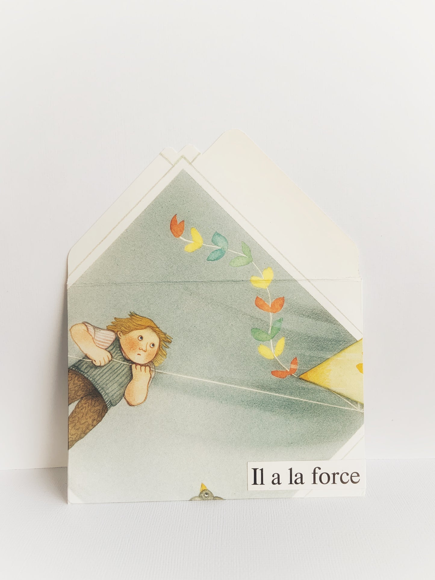 The front of a handmade envelope with the flap open showing a young boy flying a kite with a colorful tail and 'Il a la force' at the bottom.