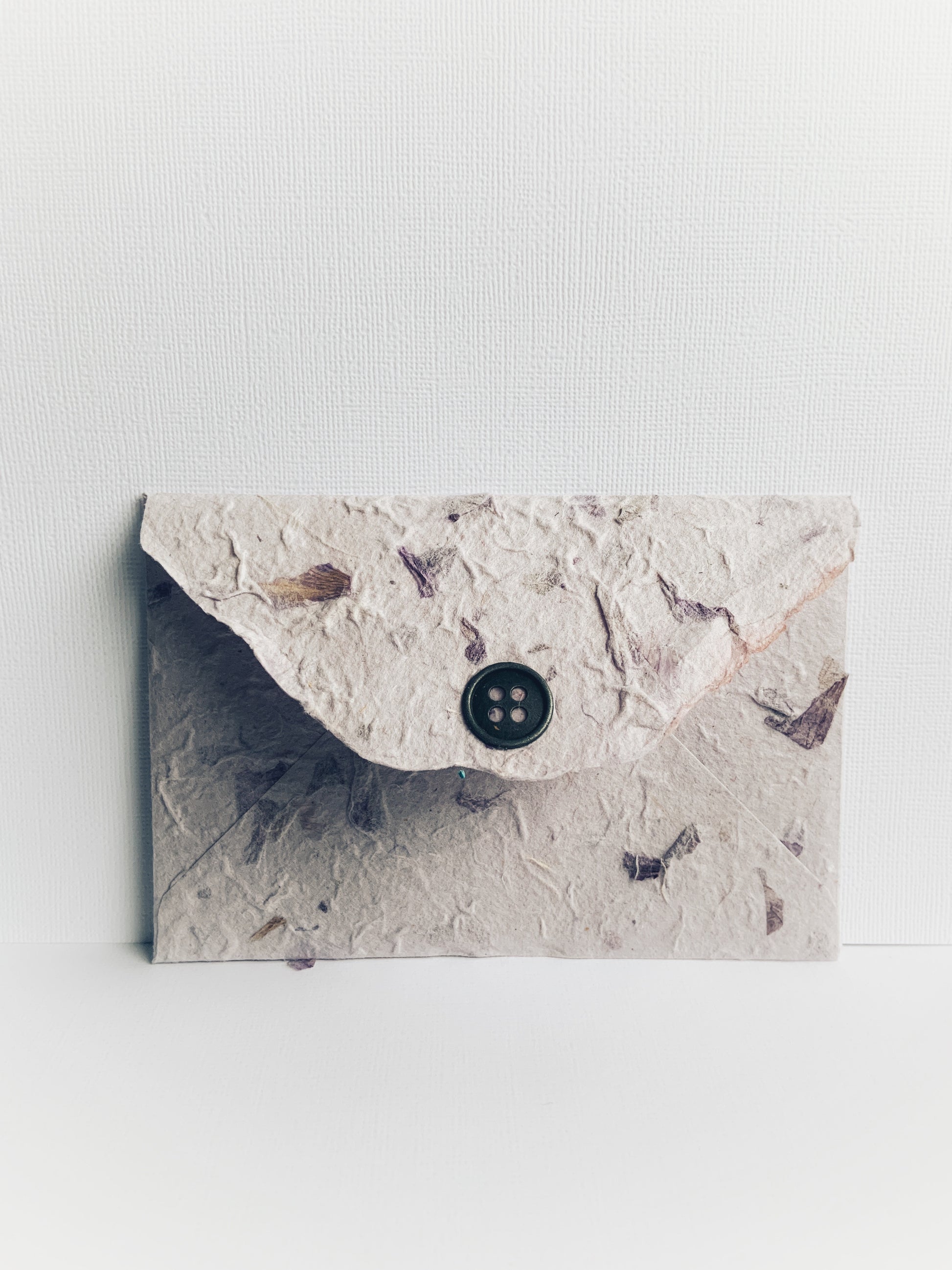 Back of a handmade envelope made from mauve handmade paper from Thailand showing detail on flap and decorative metal button. Small pieces of dried flowers add to the paper's texture.