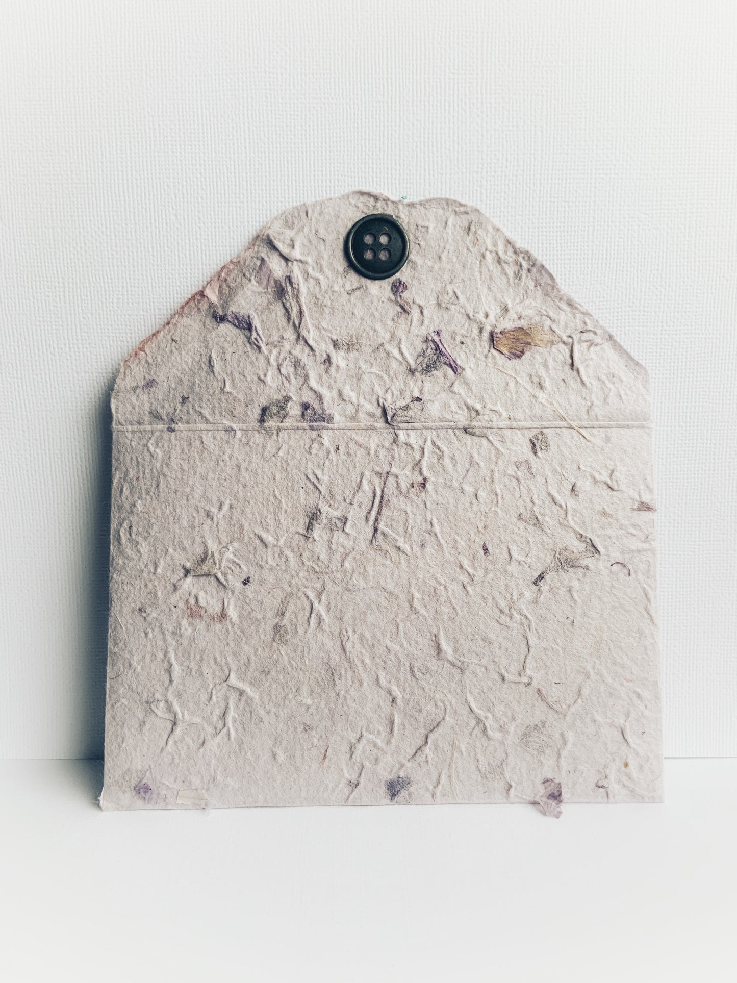 Front of a handmade envelope made from mauve handmade paper from Thailand showing detail on flap and decorative metal button. Small pieces of dried flowers add to the paper's texture.